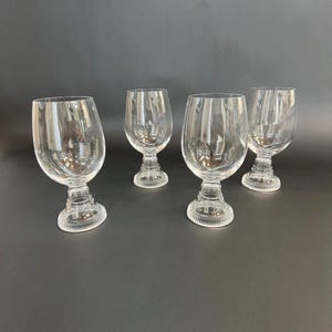 May include: Four clear glass goblets with a rounded bowl and a detailed, textured base. The stemware is transparent, allowing light to pass through, and is set against a dark background. The goblets are likely for serving beverages.
