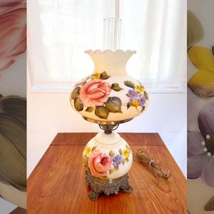 May include: A vintage table lamp with a cream-colored base and shade, adorned with hand-painted pink roses, yellow and purple flowers, and green leaves. The lamp has a clear glass chimney and a decorative metal base.