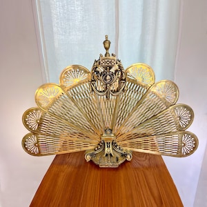 May include: Ornate brass fireplace screen with a fan-shaped design. The screen features intricate cut-out patterns and a decorative top with a detailed emblem. The brass has a warm, golden hue, and the screen is set on a wooden surface.