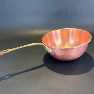 May include: A round copper saucepan with a polished finish and a long, curved brass handle. The handle is attached to the side of the bowl with two visible metal rivets. The interior of the pan is smooth and reflects light, showing a warm metallic tone.