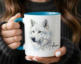 Op de afbeelding: Witte keramische mok met een blauw interieur en handvat, met een aquarel illustratie van een witte wolf. De wolf heeft gele ogen en is gesigneerd "Adam" in cursief. De mok wordt door een persoon vastgehouden.