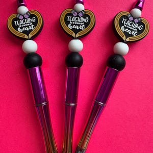 May include: Three pens with a purple and gold finish. Each pen has a heart-shaped charm with the text "TEACHING is a work of heart". The charms are black with yellow and white accents. The pens have black, white, and purple beads.