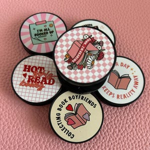Pode incluir: Uma cole&ccedil;&atilde;o de suportes redondos para telem&oacute;veis com v&aacute;rios designs e texto. Um diz "I'M ALL BOOKED UP", outro "HOT girls READ books", e outro "COLLECTING BOOK BOYFRIENDS". Outros t&ecirc;m desenhos de livros e flores.