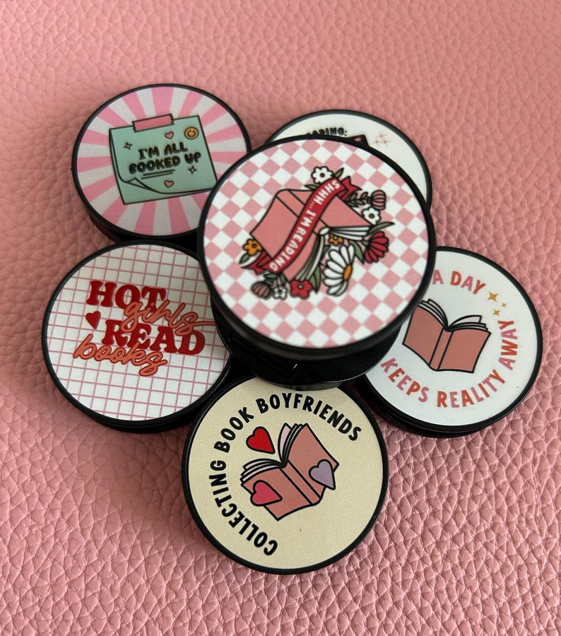 Pode incluir: Uma cole&ccedil;&atilde;o de suportes redondos para telem&oacute;veis com v&aacute;rios designs e texto. Um diz "I'M ALL BOOKED UP", outro "HOT girls READ books", e outro "COLLECTING BOOK BOYFRIENDS". Outros t&ecirc;m desenhos de livros e flores.