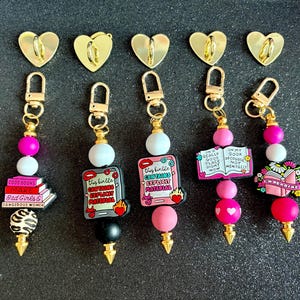 May include: A selection of keyrings with gold-tone metal fixings and decorative beads. Each keyring has a unique charm with phrases such as "Good Books Make Bad Girls" and "This Kindle Contains Explicit Material". The charms are in pink, white, and black.