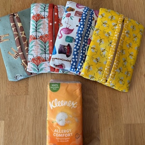 May include: A selection of fabric tissue holders with diverse patterns, such as giraffes, floral motifs, stars, flamingos, and bees. A packet of Kleenex Allergy Comfort tissues is also visible. The holders are designed to fit a standard tissue pack.