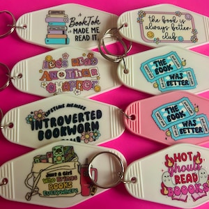May include: Ten white and pink keychains with different book-themed designs. The keychains feature phrases like "BookTok Made Me Read It", "Let's Read Another Chapter", "Introverted Bookworm", "Just a Girl Who Brings Books Everywhere", "The Book Was Better", and "Hot Ghouls Read Books".