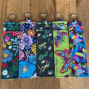 May include: Five fabric keychains with metal rings, each displaying a unique pattern. Designs include floral, butterfly, and frog motifs in vibrant colours like green, blue, and pink. The keychains are rectangular with a loop for the ring.