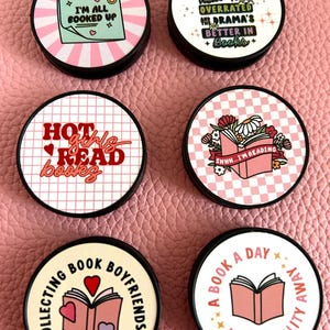 Pode incluir: Uma cole&ccedil;&atilde;o de acess&oacute;rios redondos, pretos e brancos com v&aacute;rios designs com tema de livros. Os designs incluem frases como "Estou toda reservada", "Garotas quentes leem livros" e "Um livro por dia mant&eacute;m a realidade longe". Os acess&oacute;rios t&ecirc;m um esquema de cores rosa e branco.