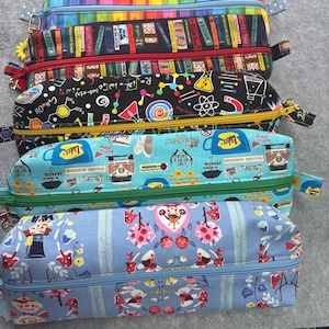 May include: A stack of six rectangular pencil cases with zipper closures. The cases feature various colorful patterns, including pencils, books, science symbols, coffee cups, and floral designs. Each case has a different colored zipper.