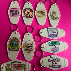 May include: Ten white motel keychains with colorful designs and inspirational messages. The keychains feature phrases like "You are worth it", "Don't let yesterday take too much of today", and "Have the courage to be disliked".
