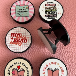 Pode incluir: Uma cole&ccedil;&atilde;o de suportes redondos para telem&oacute;vel com v&aacute;rios designs com tema de livros. Um diz "I'M ALL BOOKED UP", outro diz "READING: BECAUSE Reality's OVERRATED, DRAMA'S BETTER IN Books+". Outros suportes dizem "HOT girls READ books", "COLLECTING BOOK BOYFRIENDS" e "A BOOK A DAY KEEPS REALITY AWAY".