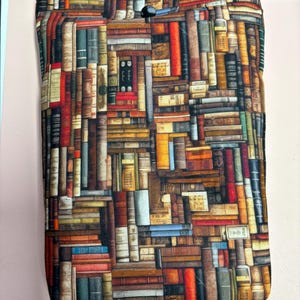May include: A rectangular book sleeve featuring a detailed print of a library filled with various books. The design includes books of different sizes and colors, with a button closure at the top. The sleeve is designed to protect books and is made of fabric.