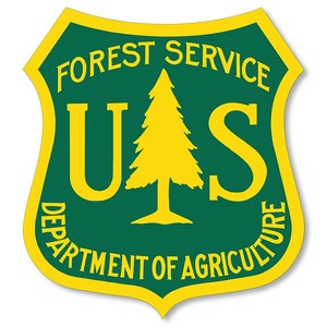 Op de afbeelding: Een groen en geel schildvormig embleem met de tekst "FOREST SERVICE" en "US DEPARTMENT OF AGRICULTURE" rond een gestileerde dennenboom.