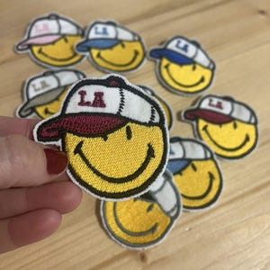 Op de afbeelding: Geborduurde patches met een lachend gezicht met een baseballpet met de letters "LA". De patches zijn geel met witte, rode en blauwe accenten op de petten. Op een houten oppervlak.