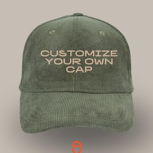 Può includere: Un cappellino in velluto a coste verde salvia con la scritta "CUSTOMIZE YOUR OWN CAP" ricamata in un colore più chiaro. Il cappellino ha una visiera curva e una corona strutturata. Il logo del marchio "vincent daimon" è visualizzato in arancione sotto.