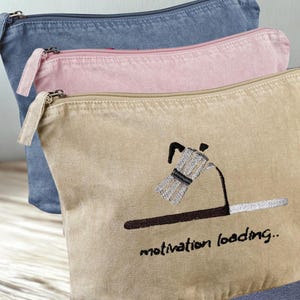 Può includere: Tre pochette con cerniera in tela, blu denim, rosa chiaro e beige. La pochette beige presenta un ricamo di una caffettiera con la scritta "motivation loading..". Ogni pochette ha una cerniera e un tirante abbinati.
