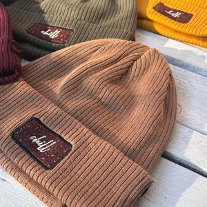 May include: A selection of ribbed knit beanies in shades of brown, olive green, and yellow. Each beanie has a rectangular patch with the word "chill" in white script. The beanies are displayed on a light-coloured wooden surface.