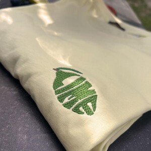 Può includere: Una maglietta giallo pallido piegata con un design a foglia ricamata verde. La forma della foglia contiene la parola "LAFE" in filo verde. La maglietta è realizzata in un materiale morbido ed è piegata.