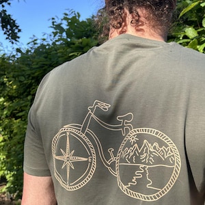 Könnte beinhalten: Olivgrünes T-Shirt mit einem Fahrrad-Design. Das Vorderrad zeigt eine Windrose und das Hinterrad eine Berglandschaft. Die Fahrradgrafik ist in einem hellbraunen Farbton gehalten.
