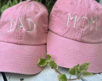 May include: Two pink baseball caps. One cap has the word "DAD" embroidered in white, and the other has "MOM". The caps are made of a soft fabric and have a curved brim. A sprig of green leaves is in the foreground.