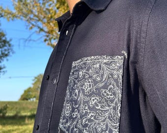 Può includere: Una camicia blu navy con tasca floreale. La tasca presenta un motivo floreale bianco su sfondo blu navy. La camicia ha cuciture e bottoni bianchi. Anche il colletto è blu navy.