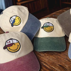 Può includere: Cappellini da baseball in vari colori, tra cui crema, bordeaux e blu navy. Ogni cappellino presenta una toppa con una faccina sorridente con le lettere "LA" sulla visiera. I cappellini hanno un classico design a sei pannelli e una visiera curva.