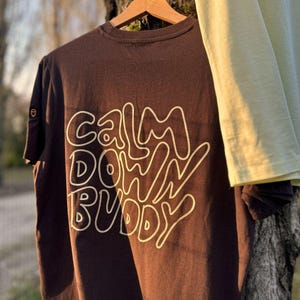 Può includere: Una maglietta marrone con la scritta "Calm Down Buddy" in un carattere ondulato bianco sporco. La maglietta è appesa a una gruccia di legno, con un'altra maglietta verde chiaro parzialmente visibile. Girocollo e maniche corte.