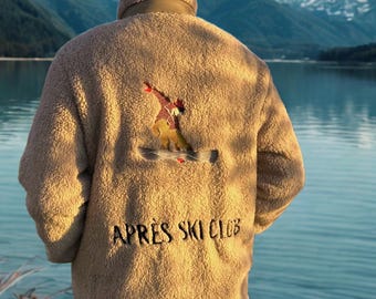 May include: A tan fleece jacket with a high collar and embroidered skier design on the back. The jacket features the text "APRES SKI CLUB" in black. The background shows a lake and snow-capped mountains.