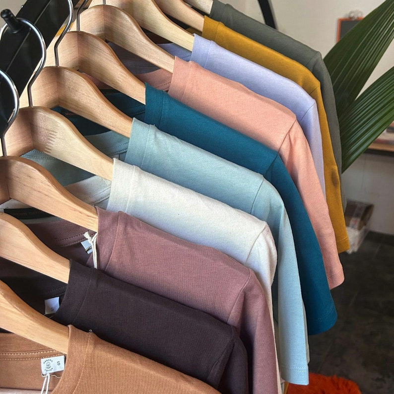 May include: A stack of plain t-shirts in various colors, including brown, beige, teal, light blue, white, and gray, hanging on wooden hangers.