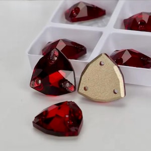 Könnte beinhalten: Eine Sammlung tiefroter, dreieckig facettierter Kristalle mit drei kleinen Löchern zum Annähen. Die Kristalle haben eine polierte, reflektierende Oberfläche und eine flache, goldfarbene Rückseite und sind in einem weißen Kunststoff-Sortierkasten angeordnet.