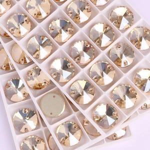 May include: Close-up of a tray filled with numerous round, faceted, golden-colored rhinestones. Each rhinestone is set in a white plastic compartment, creating a grid-like pattern. The rhinestones have a reflective, sparkling appearance, ideal for crafting and embellishment.
