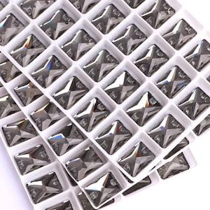 May include: Close-up of a tray filled with numerous gray, star-shaped rhinestones. Each rhinestone is set within a white, square compartment, creating a grid-like pattern. The rhinestones have multiple facets, reflecting light and adding a sparkling effect.