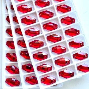 May include: A close-up of a white tray filled with rows of red, faceted rhinestones. Each gem is rectangular with two holes and a flat back. The rhinestones are a bright, transparent red colour.