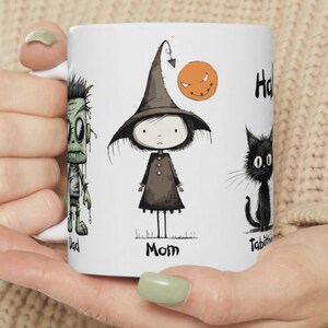 May include: White ceramic mug featuring Halloween-themed illustrations. The design includes a zombie labeled "Dad", a witch labeled "Mom", a smiling jack-o'-lantern, and a black cat labeled "Tabitha". The mug is held by a person.