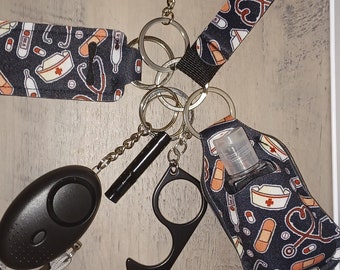 May include: A collection of accessories on keychains, including a black pom-pom, a wristlet with a medical theme, a personal safety alarm, a touch-free door opener, and a hand sanitizer holder. The wristlet features medical symbols.