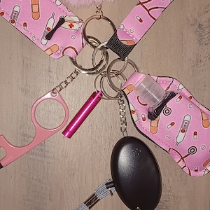 May include: A pink and white patterned keychain with a pink fuzzy pom-pom, a pink tool, a small clear bottle, a black personal alarm, and a gray lanyard.