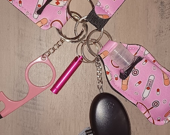 May include: A pink and white patterned keychain with a pink fuzzy pom-pom, a pink tool, a small clear bottle, a black personal alarm, and a gray lanyard.