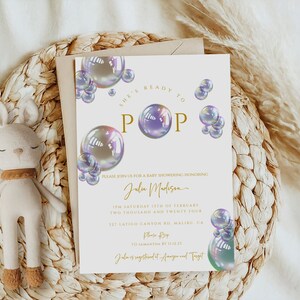 May include: A white baby shower invitation with a gold and purple theme. The invitation features a design of iridescent bubbles with the text "She's Ready to Pop" in gold lettering. The invitation includes the name "Julu Madison" and details for the baby shower.