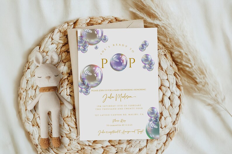 May include: A white baby shower invitation with a gold and purple theme. The invitation features a design of iridescent bubbles with the text "She's Ready to Pop" in gold lettering. The invitation includes the name "Julu Madison" and details for the baby shower.