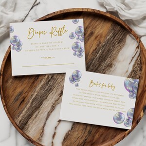 May include: Two white cards with gold text and iridescent bubble illustrations. The top card says "Diaper Raffle" and asks guests to bring a pack of diapers for a chance to win a prize. The bottom card says "Books for Baby" and asks guests to bring a book instead of a card.