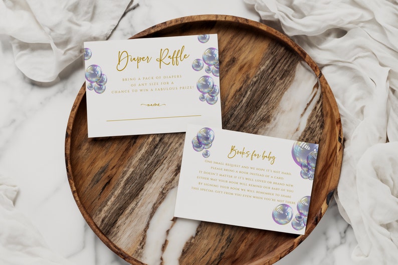 May include: Two white cards with gold text and iridescent bubble illustrations. The top card says "Diaper Raffle" and asks guests to bring a pack of diapers for a chance to win a prize. The bottom card says "Books for Baby" and asks guests to bring a book instead of a card.