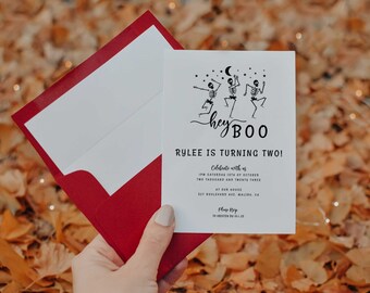 Op de afbeelding: Een witte uitnodigingskaart met een rode envelop. De kaart heeft een zwart-witte illustratie van drie skeletten die dansen met de tekst "hey BOO" en "RYLEE IS TURNING TWO!" De uitnodiging geeft details over een feest op 15 oktober 2023 om 17:00 uur.