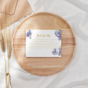 May include: A white card with gold text that reads "Books for Baby". The card is decorated with iridescent blue and purple bubbles. The text on the card reads: "One small request we have for you, it's most basic. Please bring a book instead of a gift. It doesn't matter if it's well-loved or brand new, either way your book will become our baby's too. By bringing a book, you will be helping our baby to grow. This special gift from you will be remembered for years."