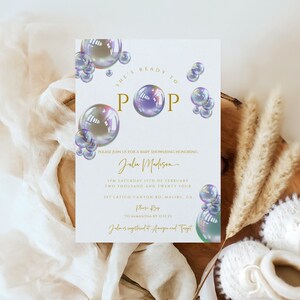 May include: A white baby shower invitation with gold and white text overlay. The text reads "She's ready to pop" with a large iridescent bubble graphic. The invitation includes the name "Julia Madison" and the date and time of the event.