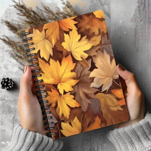 May include: A spiral-bound notebook featuring a vibrant cover design of overlapping autumn leaves in shades of golden yellow, burnt orange, and deep brown. The black metal wire binding runs along the left side of the rectangular notebook cover.