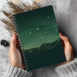 Op de afbeelding: Een spiraalgebonden notitieboek met een donker bosgroene kaft, voorzien van een illustratie van gloeiende gele sterren, zachte lichtbollen en vloeiende, golvende lijnen die een gevoel van beweging creëren op de voorzijde van de kaft.