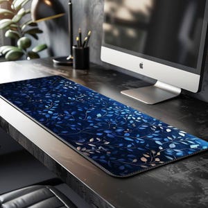 Può includere: Un tappetino da scrivania rettangolare con sfondo blu scuro e un motivo di foglie blu e bianche. Il tappetino è su una scrivania nera, accanto a un monitor del computer e un portapenne nero.