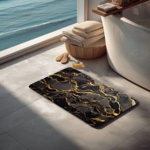 May include: A rectangular bath mat featuring a black and gray marble pattern with prominent gold veining throughout. The mat has rounded corners and a smooth surface, contrasting with the light-colored tiled floor it rests upon.