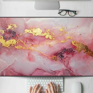 May include: A pink and gold desk mat with a marble design. The mat features a blend of pink hues with gold accents, creating a luxurious aesthetic. The design is abstract and fluid, with a glossy finish. A keyboard and mouse are visible at the bottom.